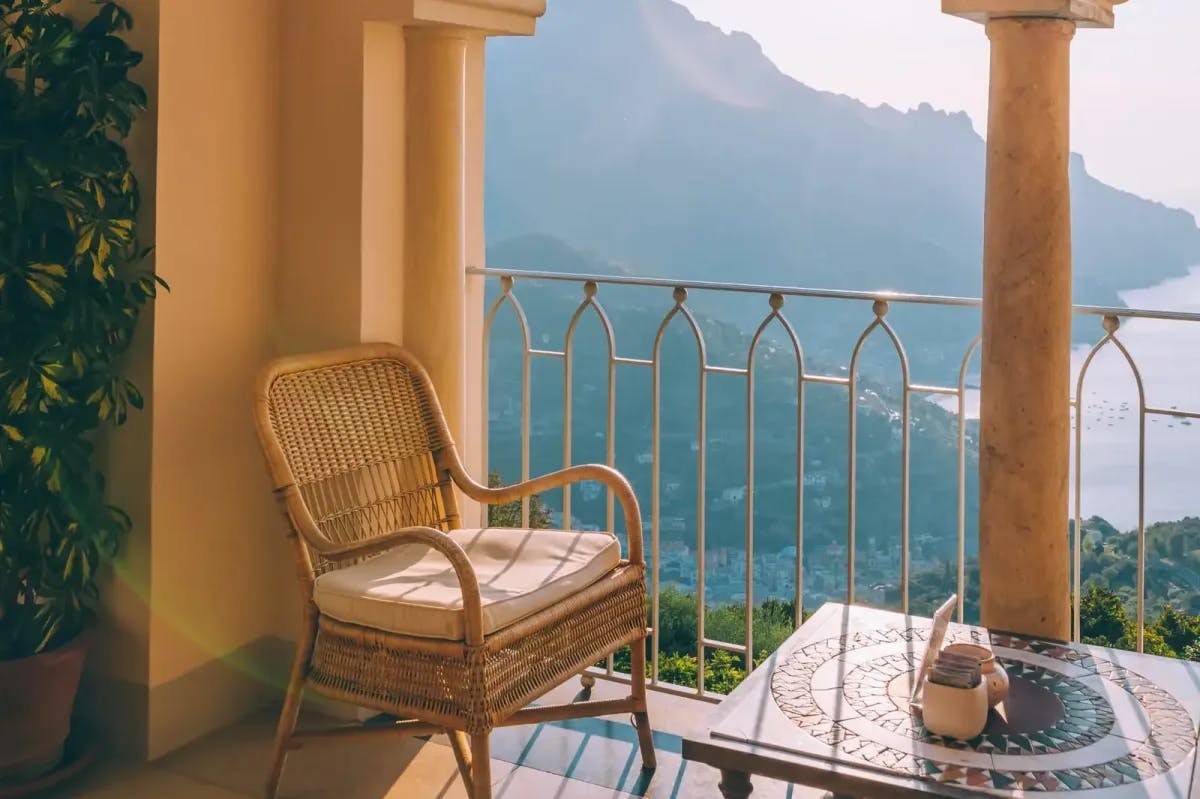 A wicker chair with a plush cushion is positioned along a balcony overlooking the Amalfi coastline